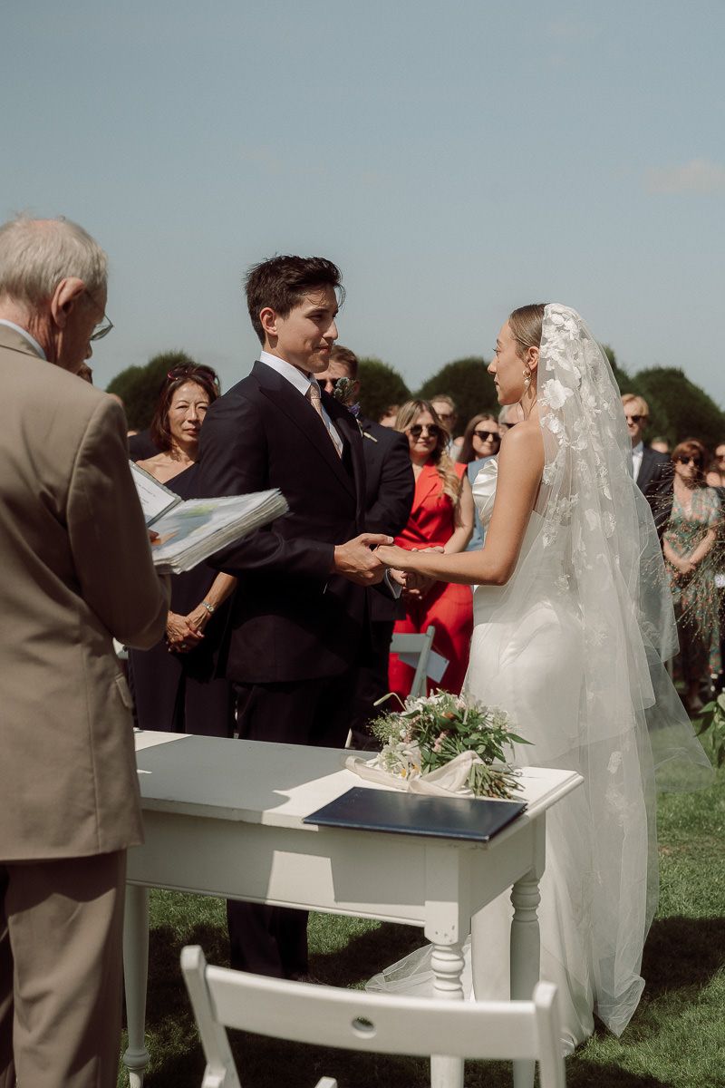 An outdoor wedding ceremony with guests gathered around as couple exchanges vows at white podium.