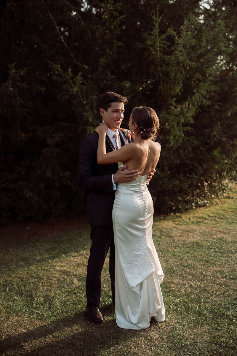 A couple in wedding attire dance together on a lawn at sunset with evergreen trees in the background.