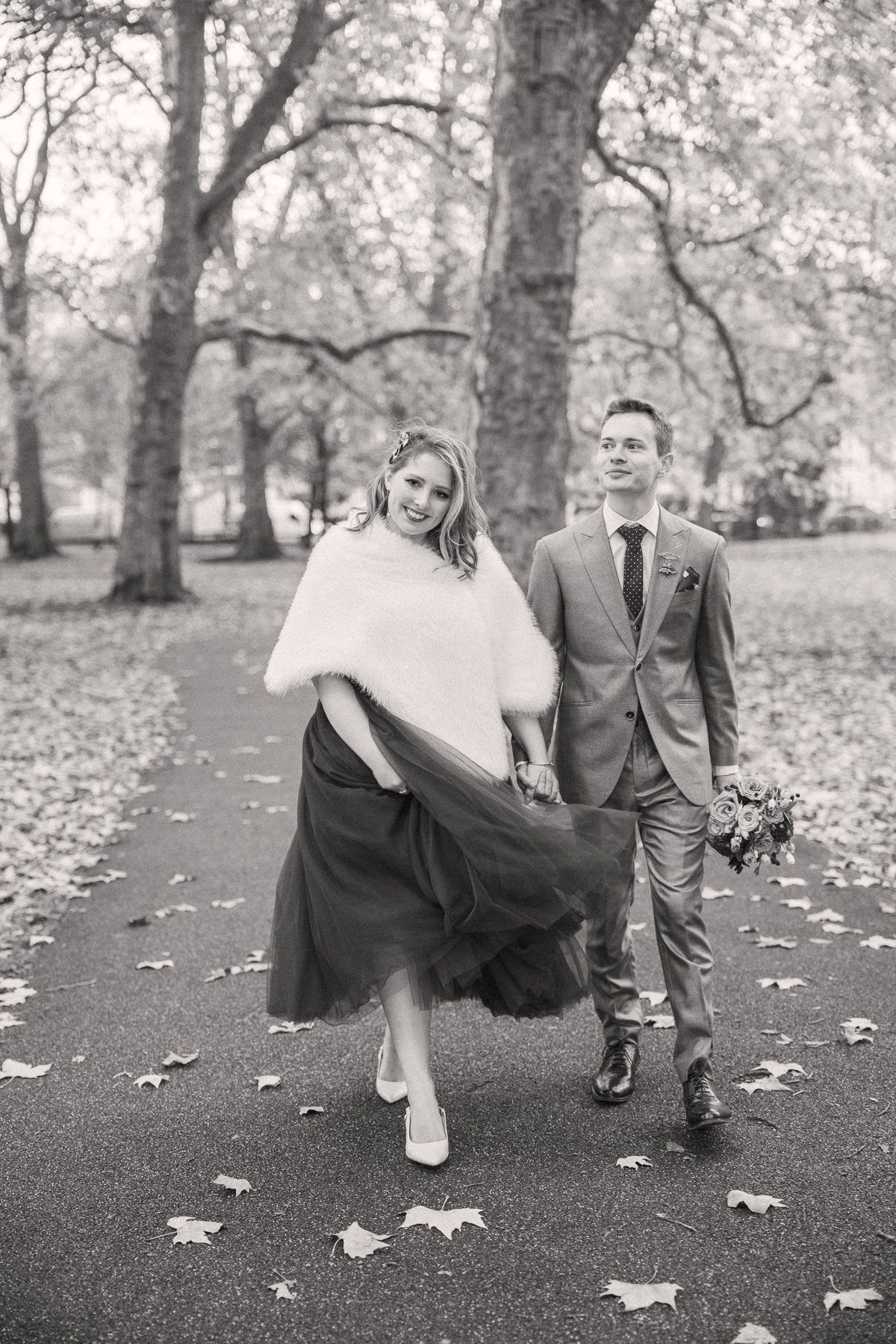 A bride and groom walk together on a path surrounded by trees in a park on their wedding day.