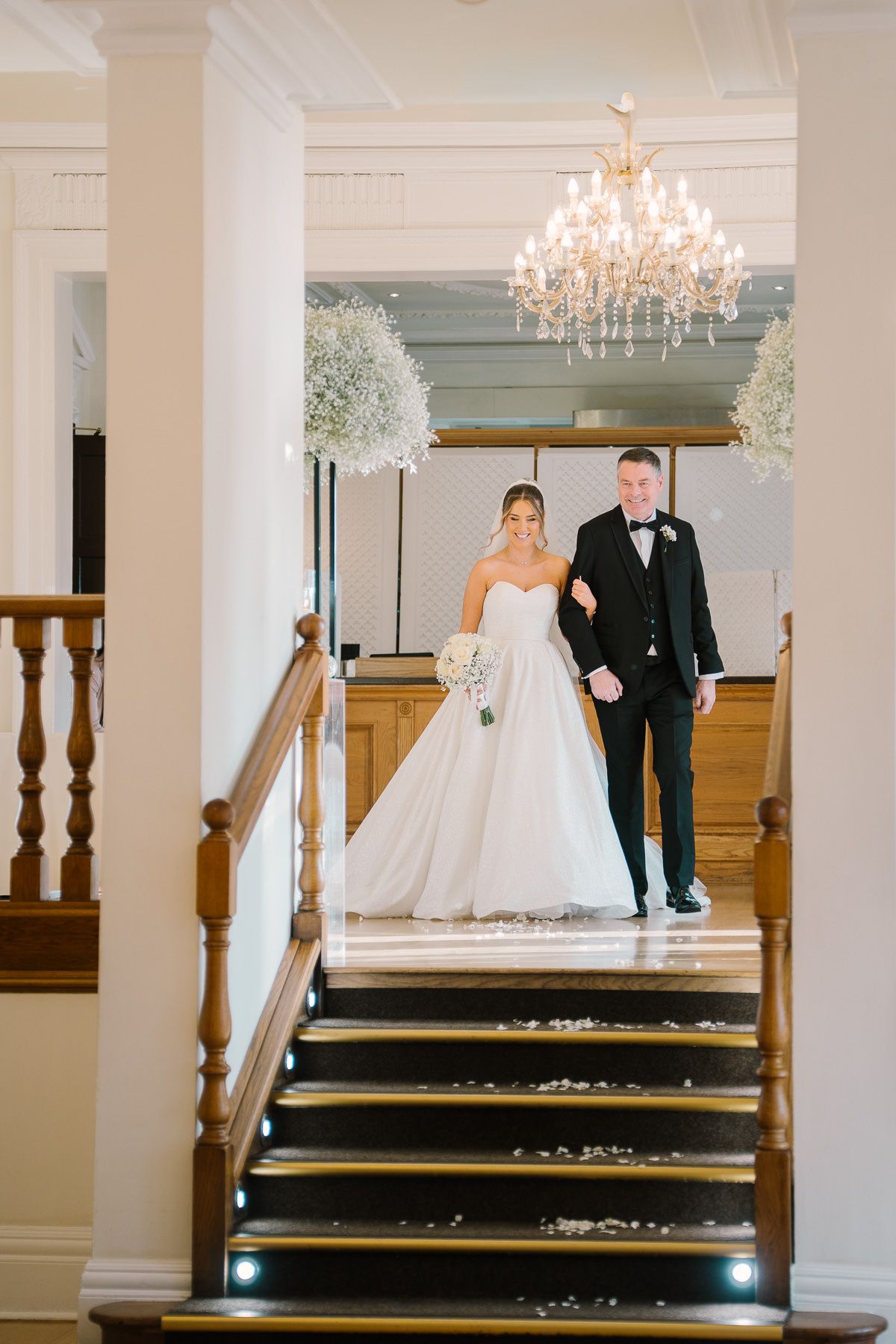A bride and groom standing on a grand staircase in a formal wedding setting, with a chandelier overhead.