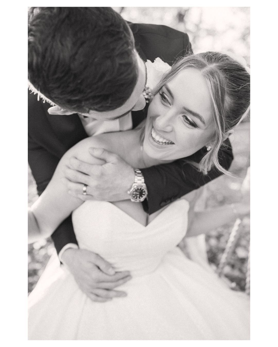 Black and white wedding photo of smiling bride in strapless gown embraced by groom in suit, sharing a romantic moment.