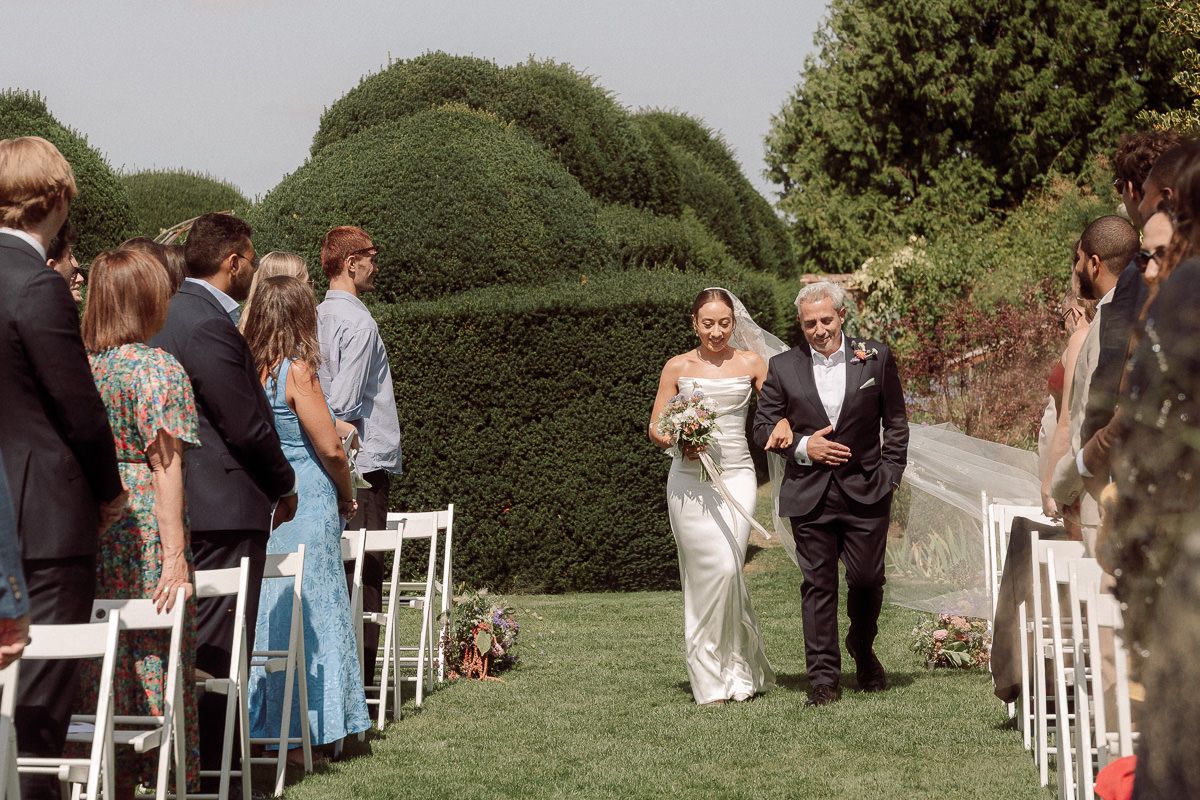 Guests gather outdoors for a wedding ceremony with white chairs arranged in rows and lush green hedges in the background.