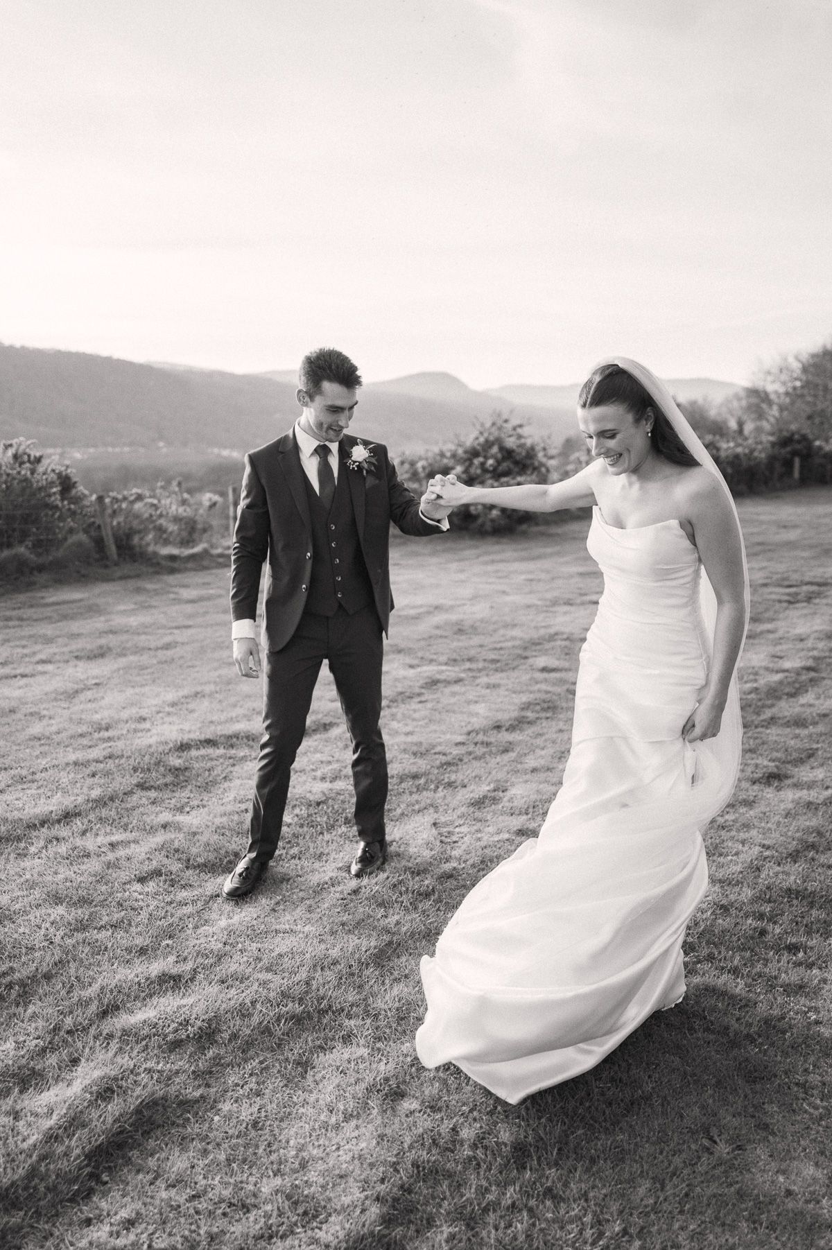 A bride in a white dress and a groom in a black suit walk together on a grassy field with mountains in the background.