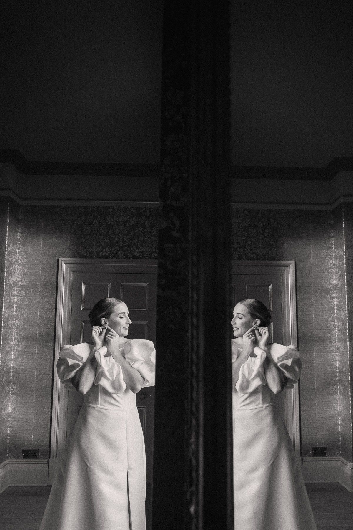 A black and white image showing a woman in a white dress standing in front of a mirrored wall.
