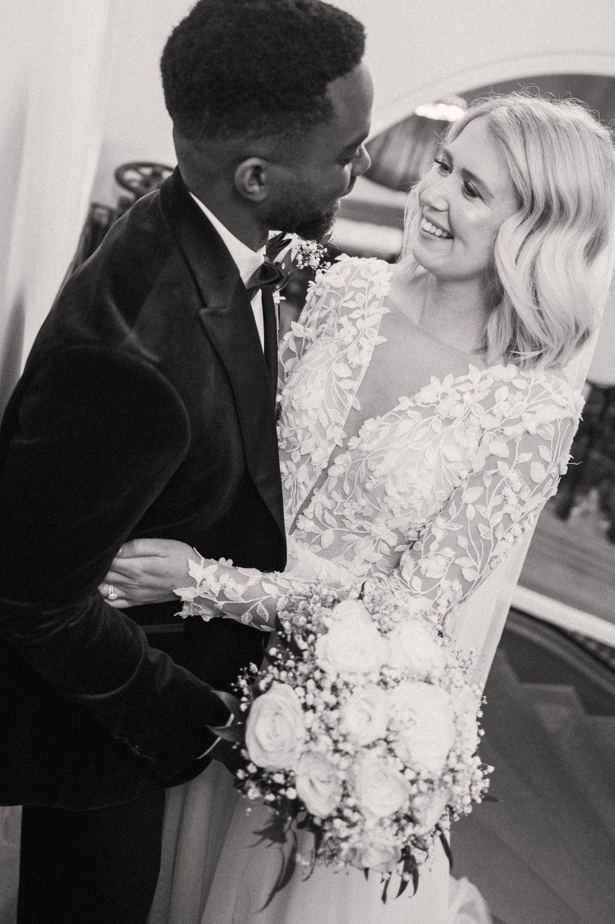 A black man in a tuxedo embracing a smiling blonde woman in a white lace wedding dress and holding a bouquet of flowers.
