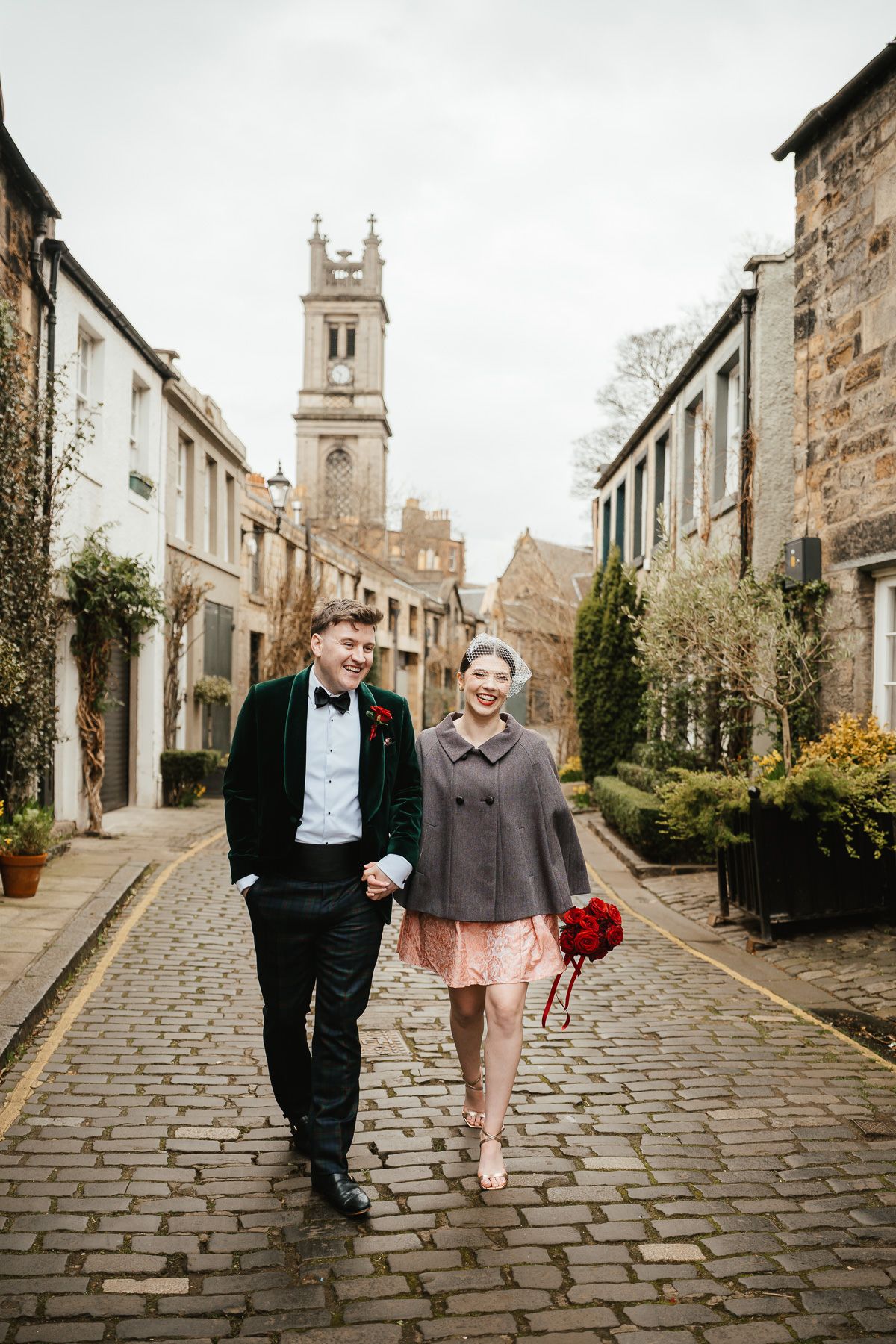 A couple walks together on a charming cobblestone street in a historic Scottish village with a church steeple visible.