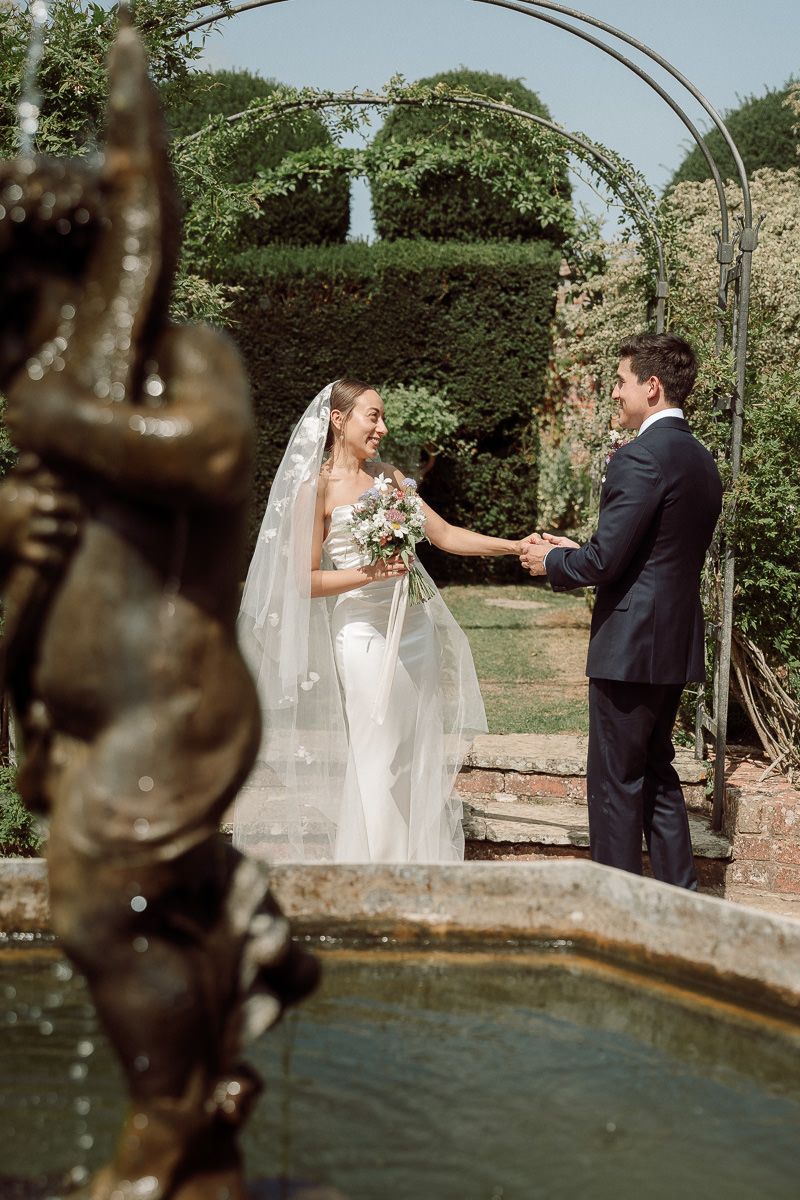 A couple shares a romantic moment under a garden arch next to a reflecting pool on their wedding day.