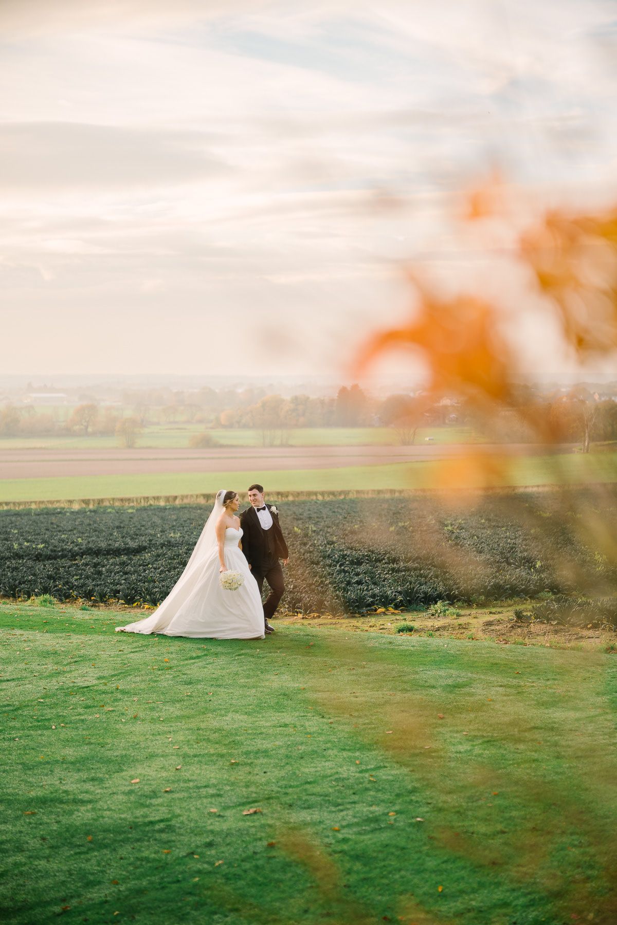 A bride and groom embrace in a scenic outdoor setting with a grassy field and autumn foliage in the background.