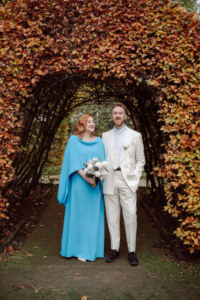 A couple in turquoise and beige attire pose together under an autumn archway of orange and brown leaves.