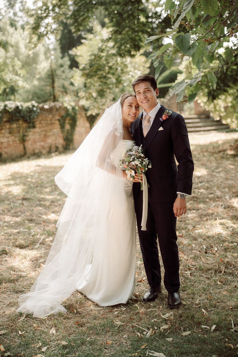 A newlywed couple poses outdoors under green trees on their wedding day, with the bride in white and groom in black suit.