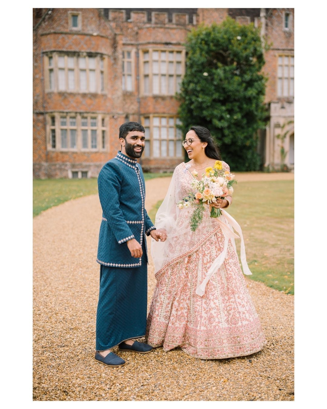 Couple in traditional Indian wedding attire posing outdoors at a grand estate, bride holding a floral bouquet.