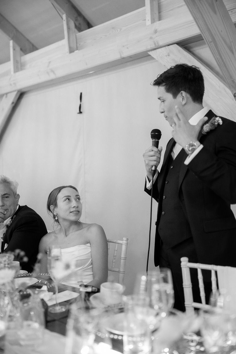 A black and white wedding reception photo showing a speech being given at a formal dinner table setting.