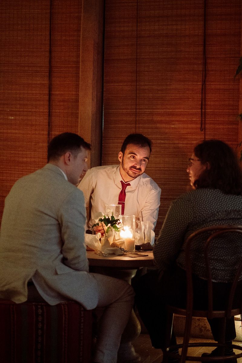 Three people having an intimate conversation at a candlelit table in a dimly lit restaurant with wooden decor.