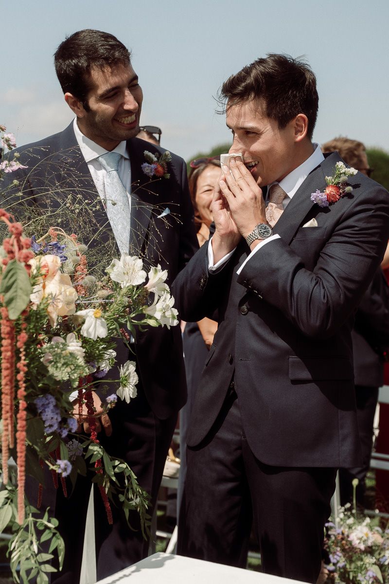 Wedding couple in formal attire laugh and embrace while holding bouquets during an outdoor celebration.