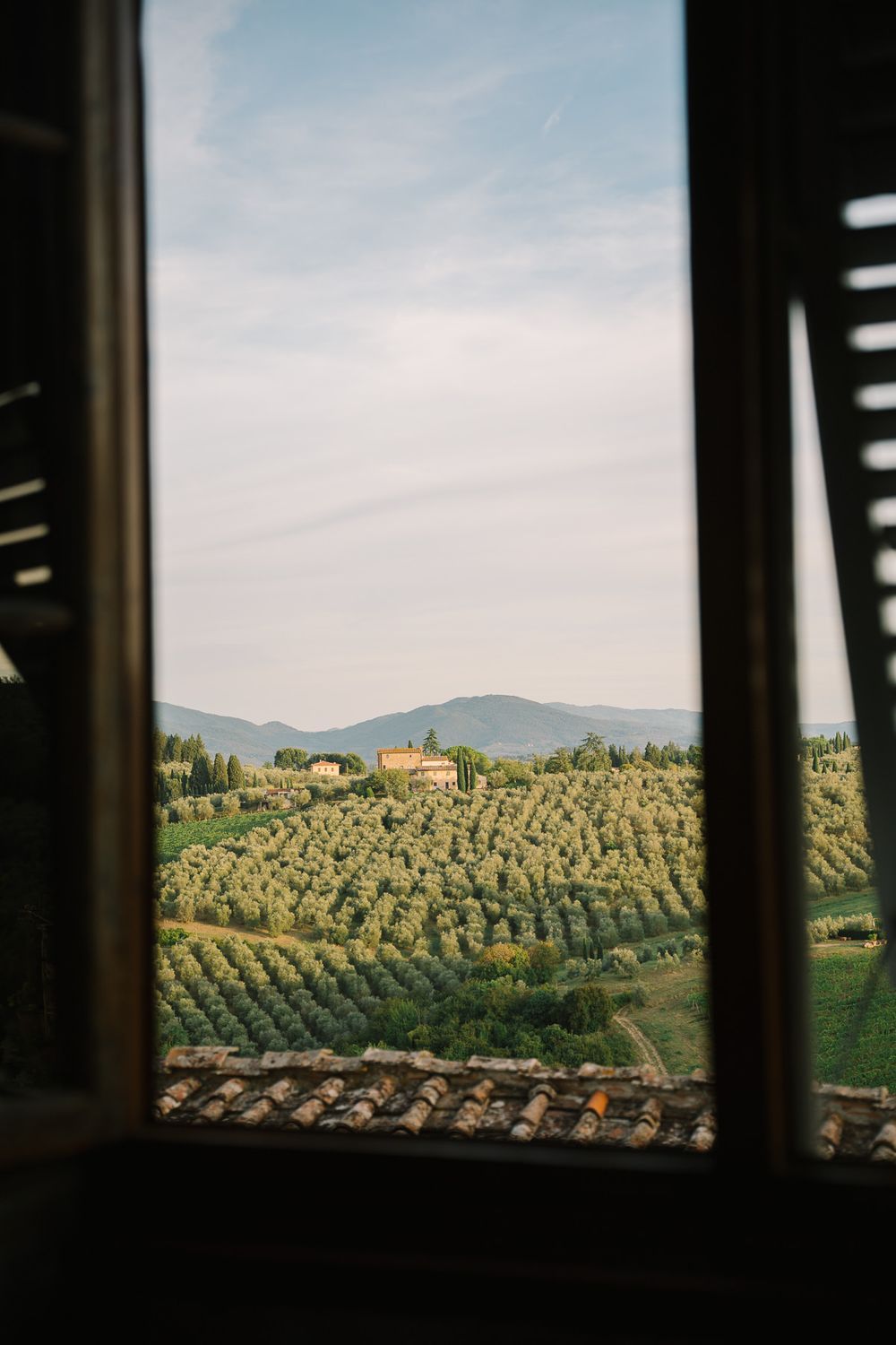 View through open window of rolling Tuscan hills with olive groves, cypress trees, and a farmhouse under a blue sky.