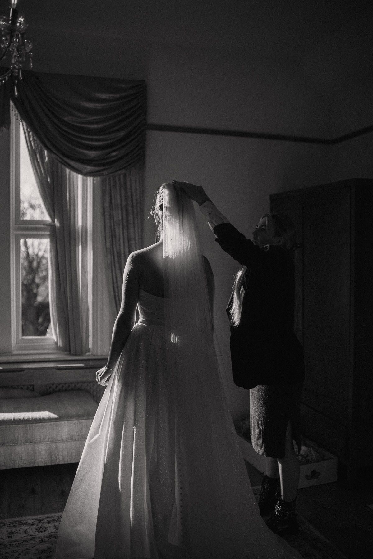 A black and white image of a bride in a long, flowing wedding dress being helped by a person, likely the groom, in a dimly lit room.