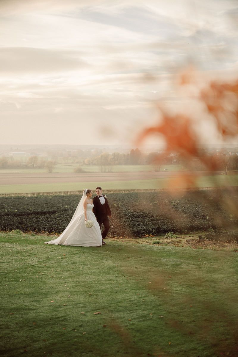 A couple in wedding attire stands together on a green hillside overlooking a scenic landscape at dusk.