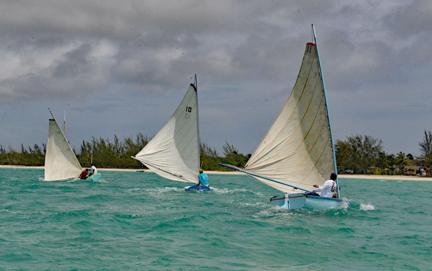 Three white sailboats racing across turquoise waters on a cloudy day.