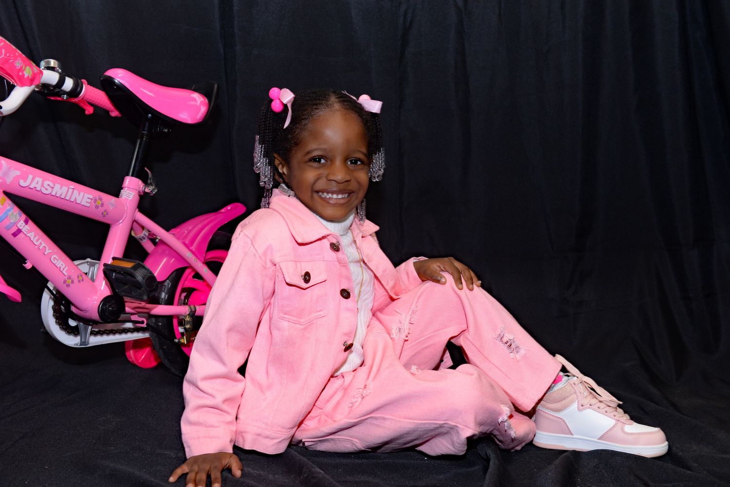 Child in bright pink outfit poses next to pink bicycle against dark background.