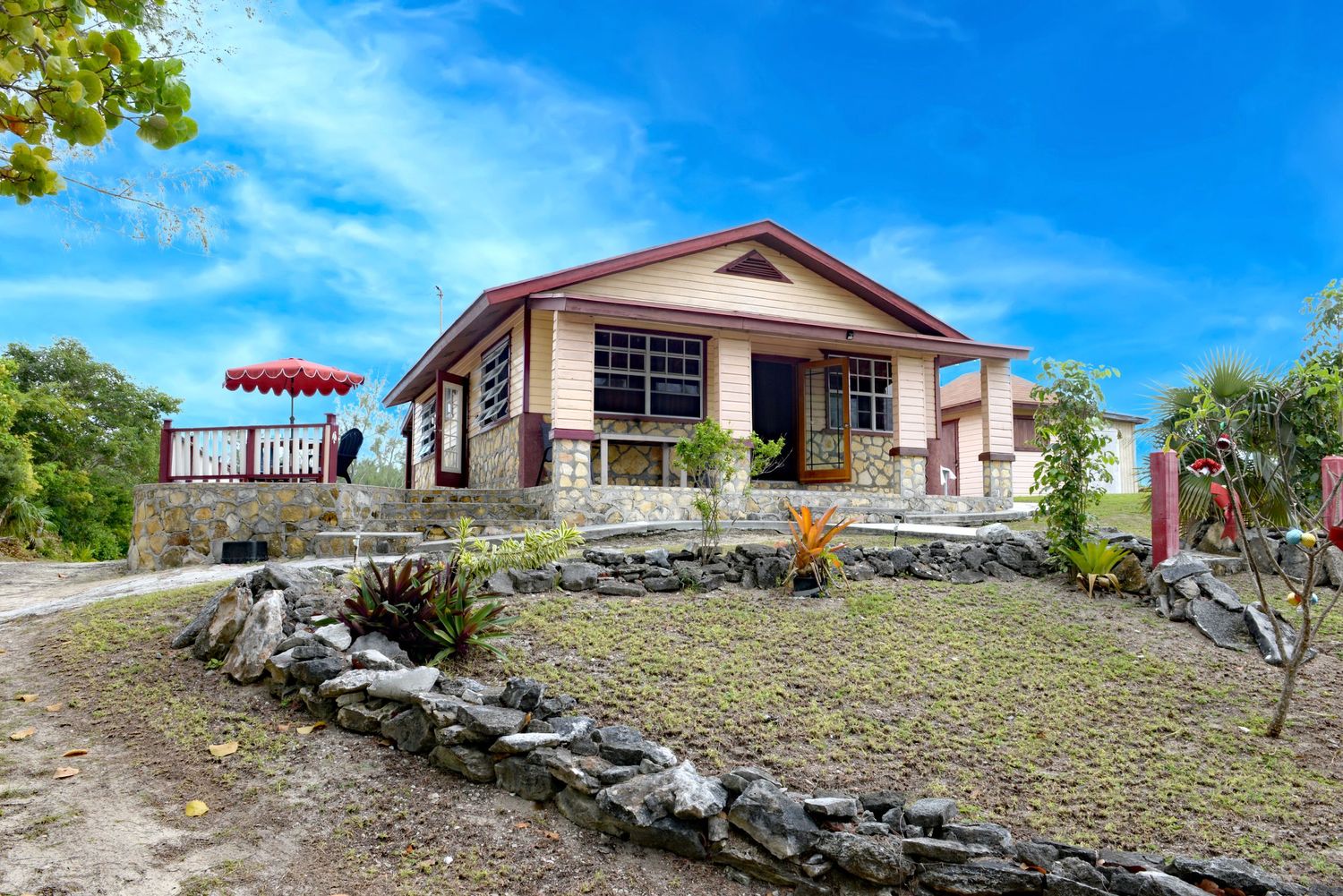 Single-story ranch house with stone wall surrounded by tropical vegetation against blue sky.