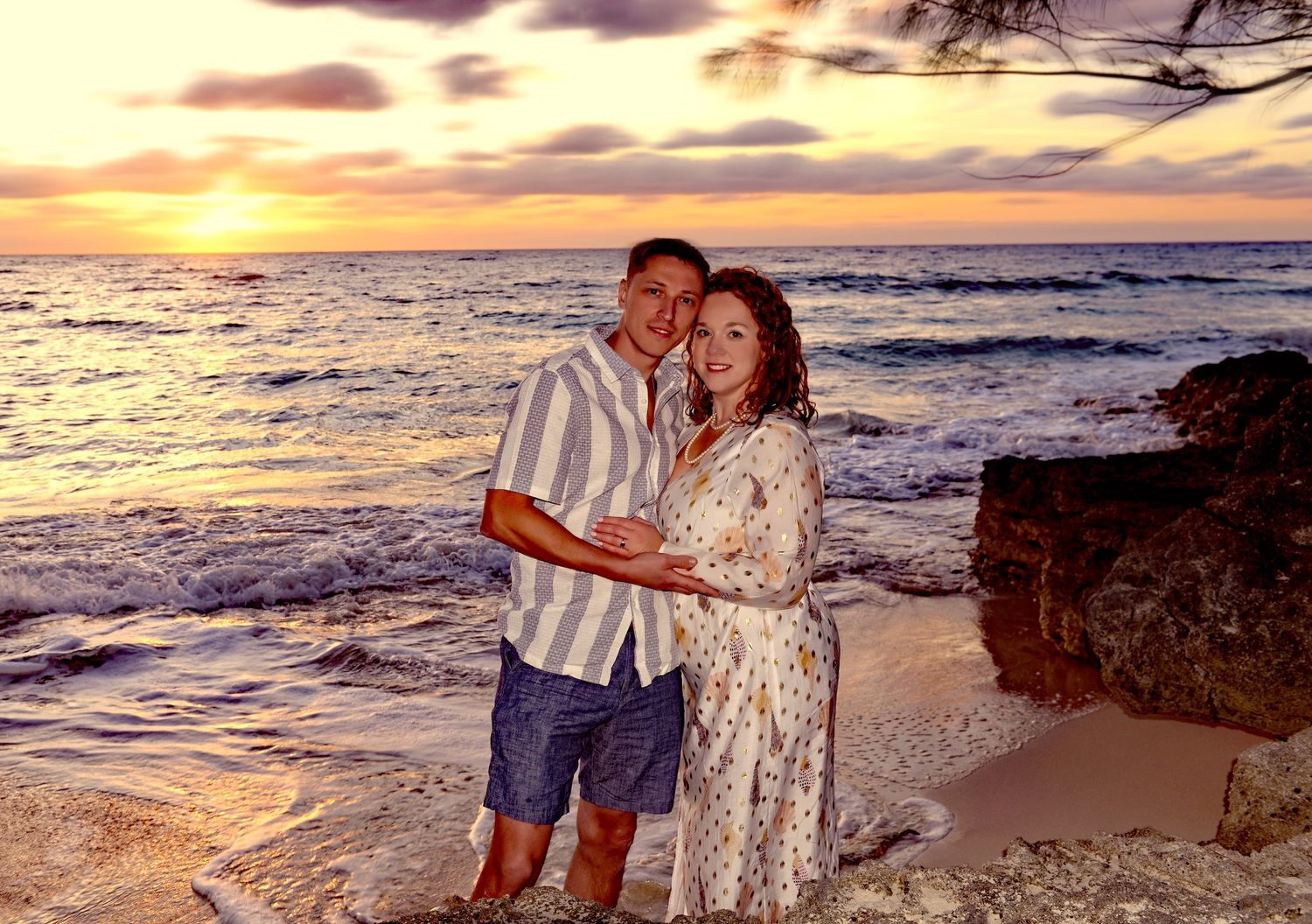 A couple shares an intimate moment on a beach at sunset with waves crashing against rocky shoreline in Hawaii.
