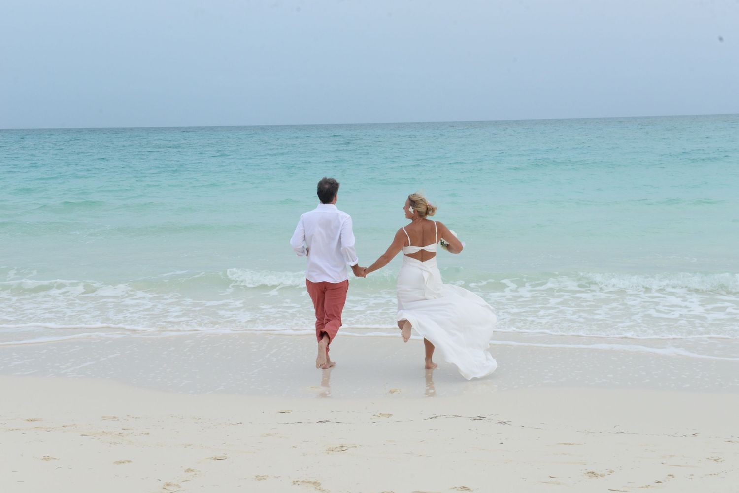 Joyful newlywed couple running along pristine beach with turquoise water in background.