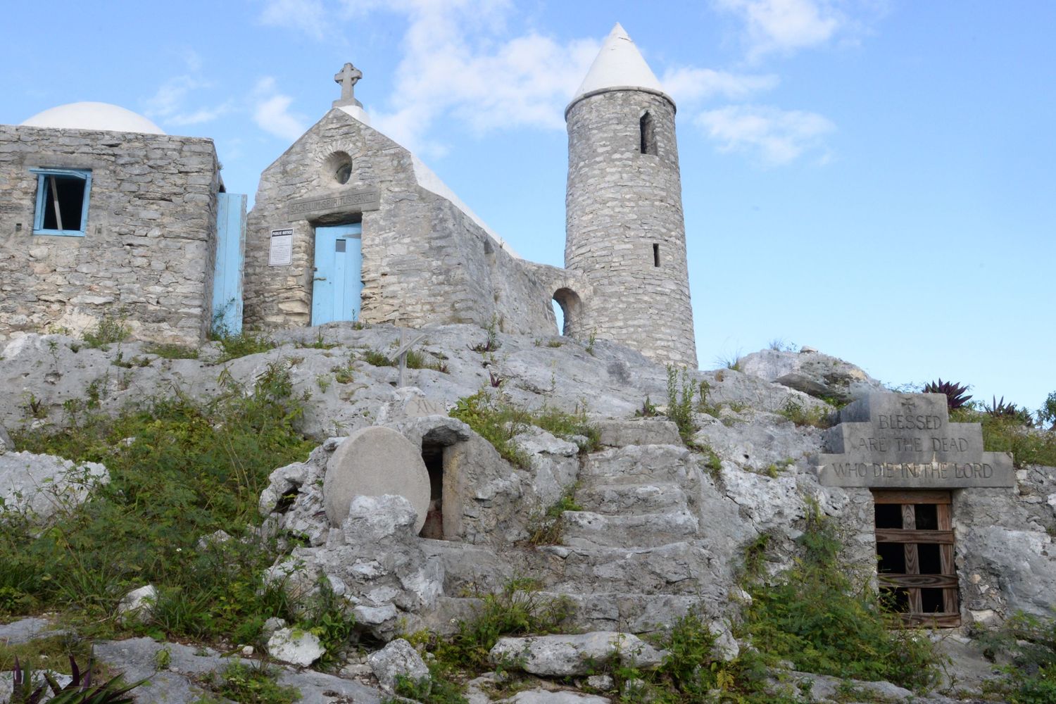 Ancient stone church with a tower and cross perched on a rocky hillside under blue skies.