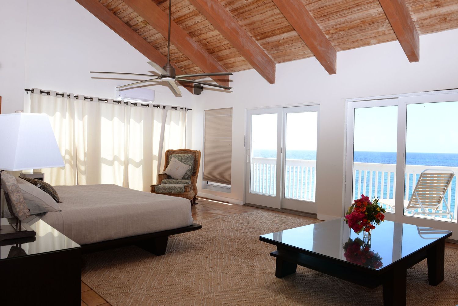 Luxurious oceanfront bedroom with exposed wooden beams, white bedding, and panoramic sea views through large windows.