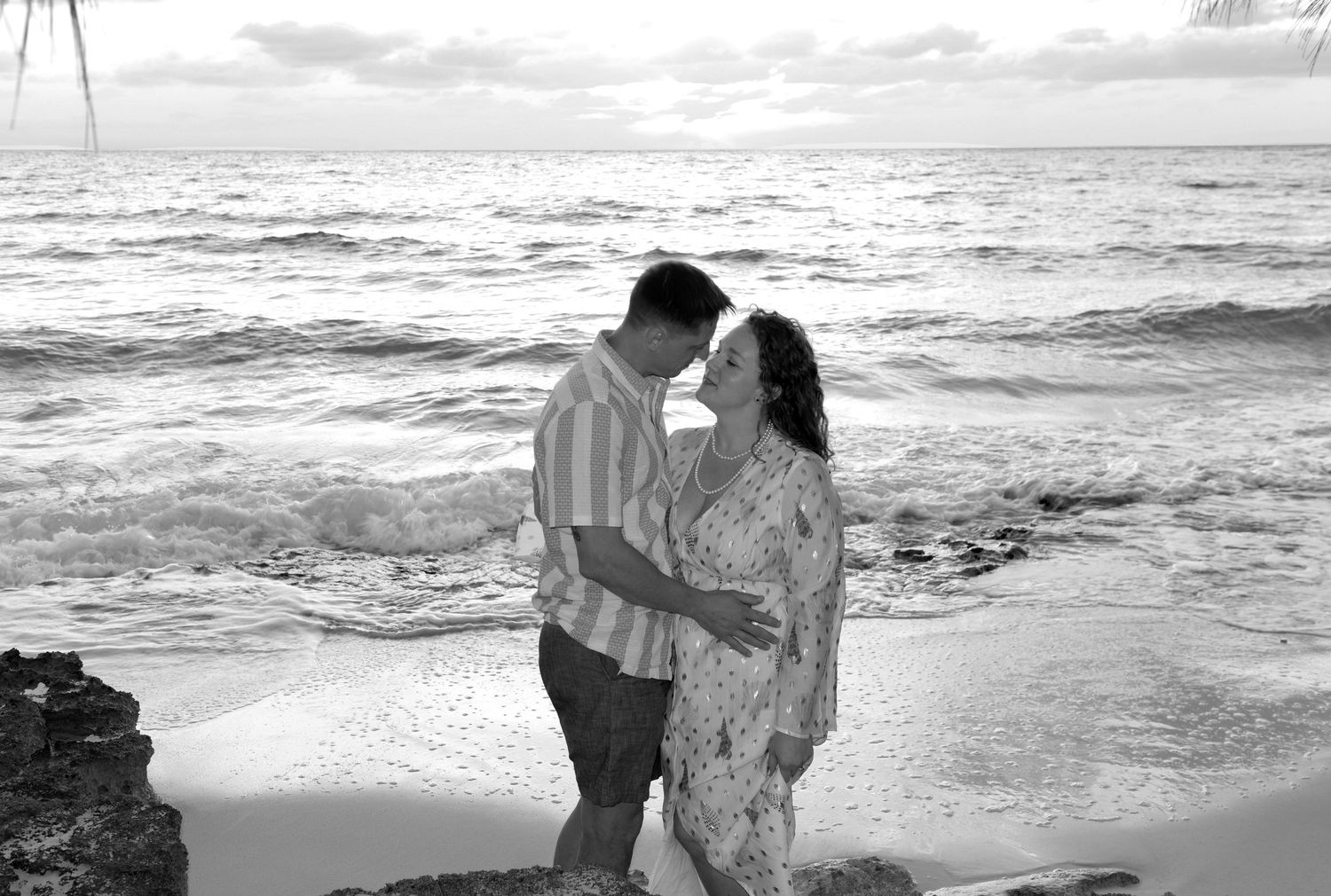 A romantic couple in black and white embraces on a sandy beach with ocean waves in the background.