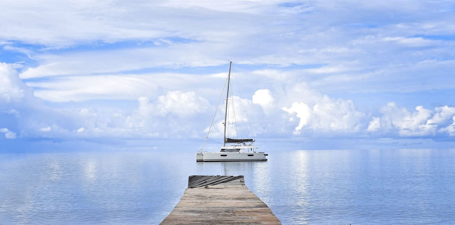A wooden pier extends into calm blue waters with a sailboat moored at the end beneath puffy white clouds.