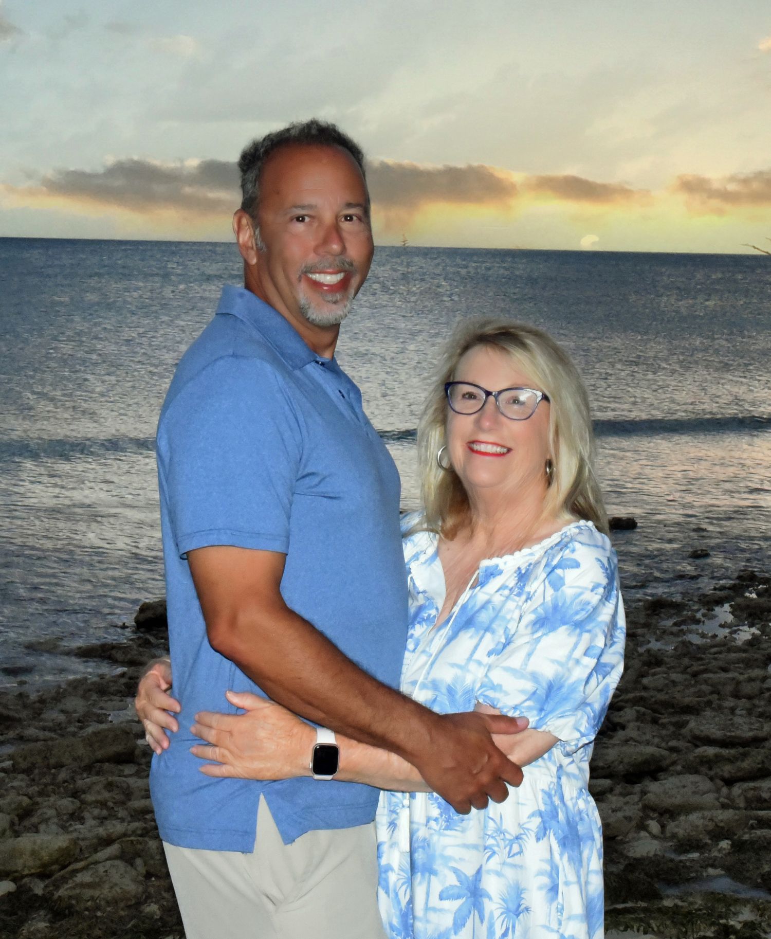 A couple embraces on a rocky beach at sunset with the ocean and glowing sky in the background.