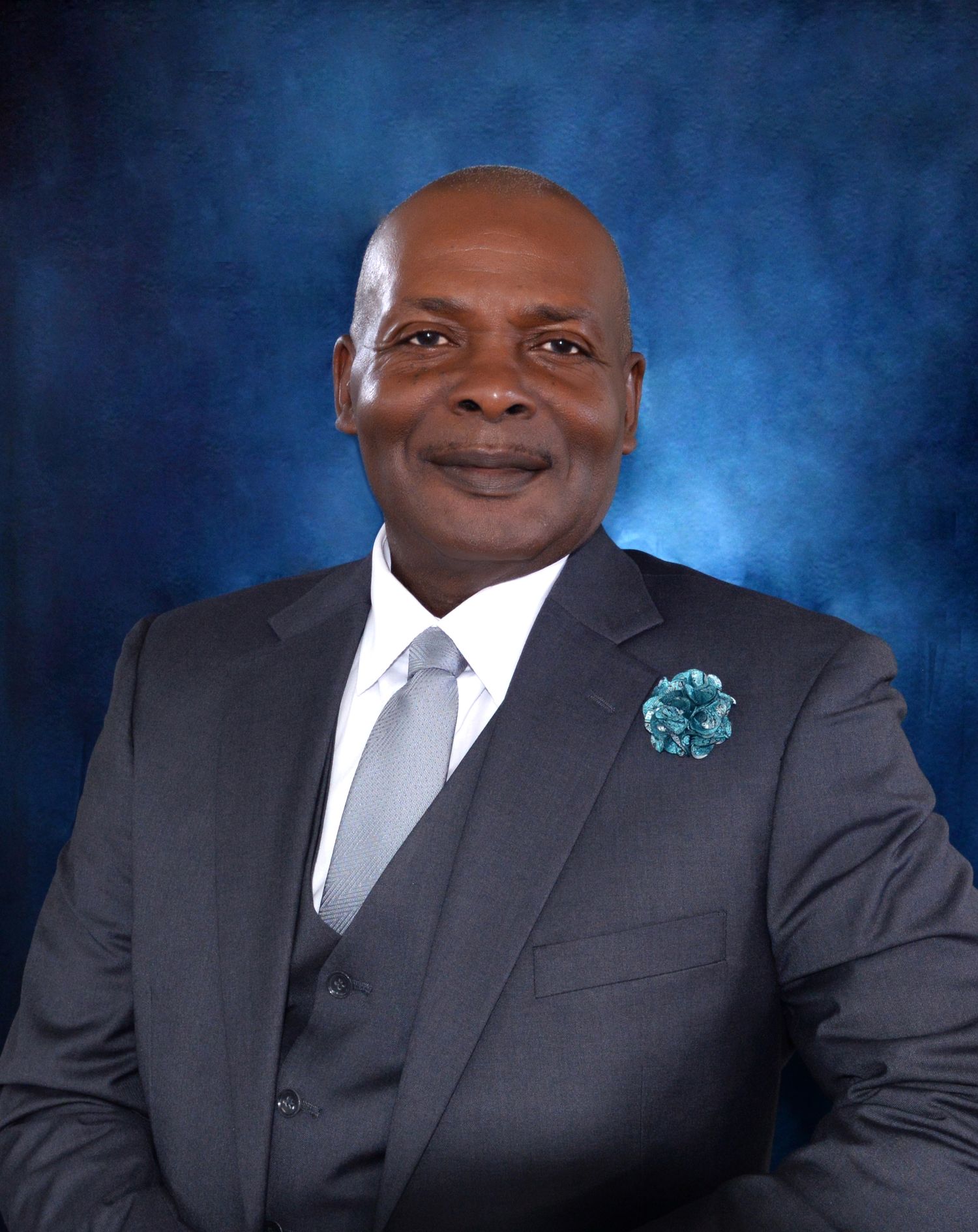 Professional portrait of businessman wearing dark suit with teal boutonniere against blue backdrop.