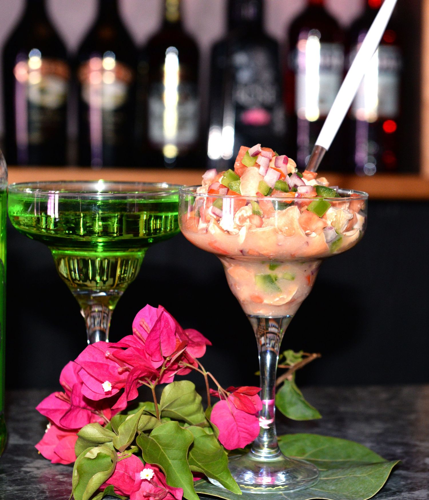 Colorful cocktails and bright pink flowers displayed at a bar with wine bottles in background.