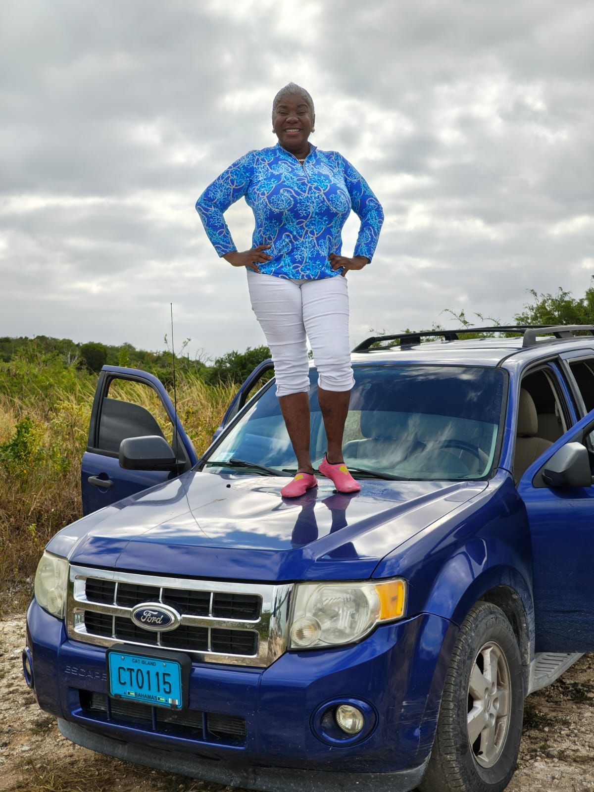 Person in a blue patterned top and white pants stands confidently on the hood of a dark blue Ford SUV on a cloudy day.