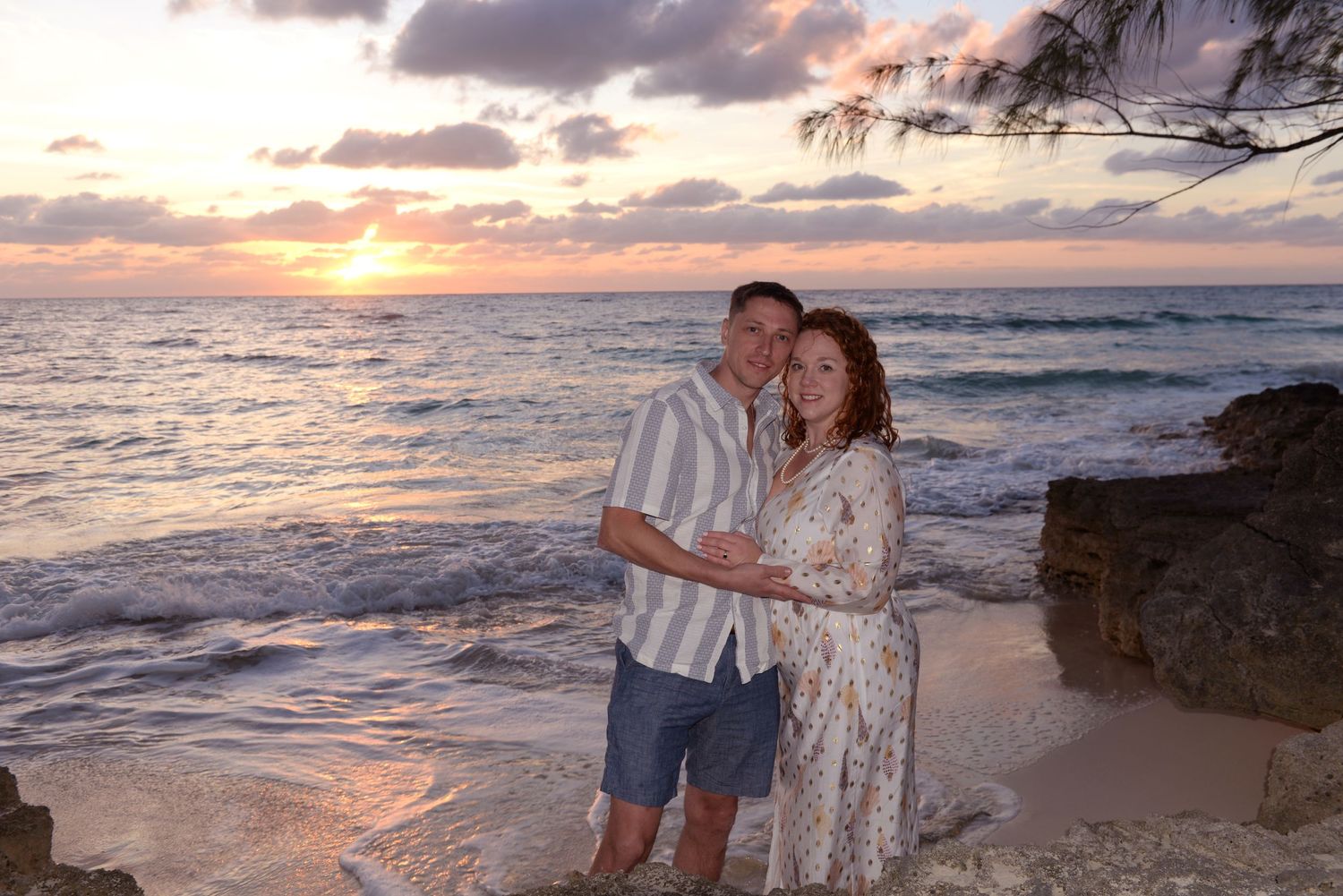 A couple stands together on a beach at sunset with ocean waves and rocky coastline in the background.