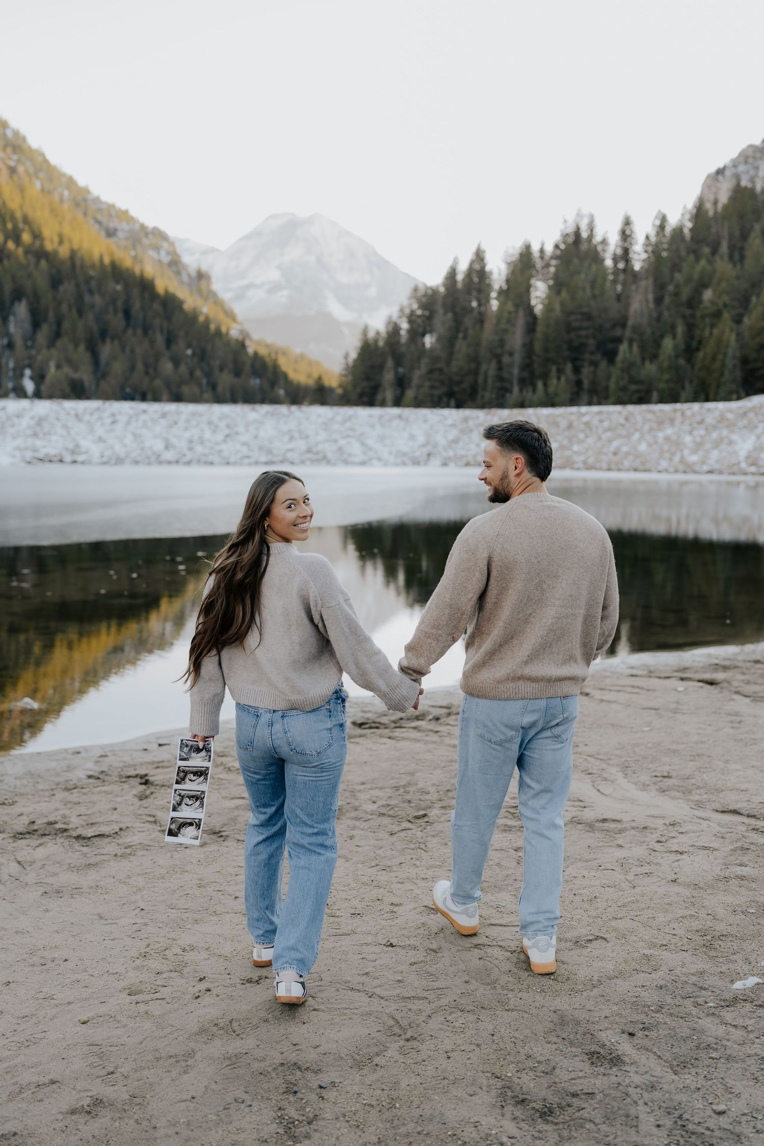 Expecting couple walking hand-in-hand along mountain lake shore, woman holding ultrasound photos.