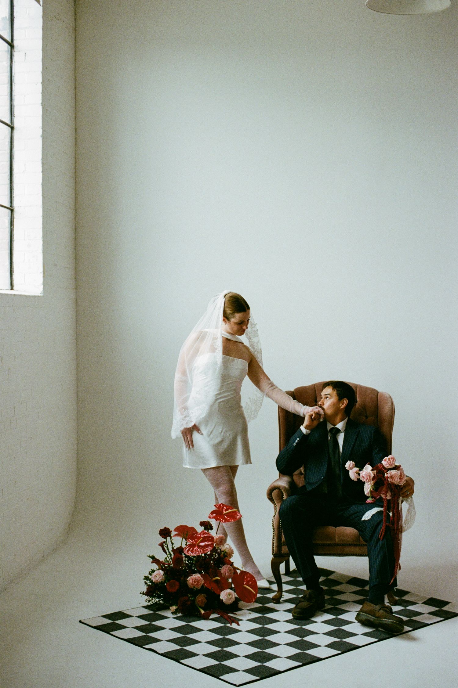 Bride in short white dress and veil stands on checkered floor holding groom's hand as he sits in leather chair.