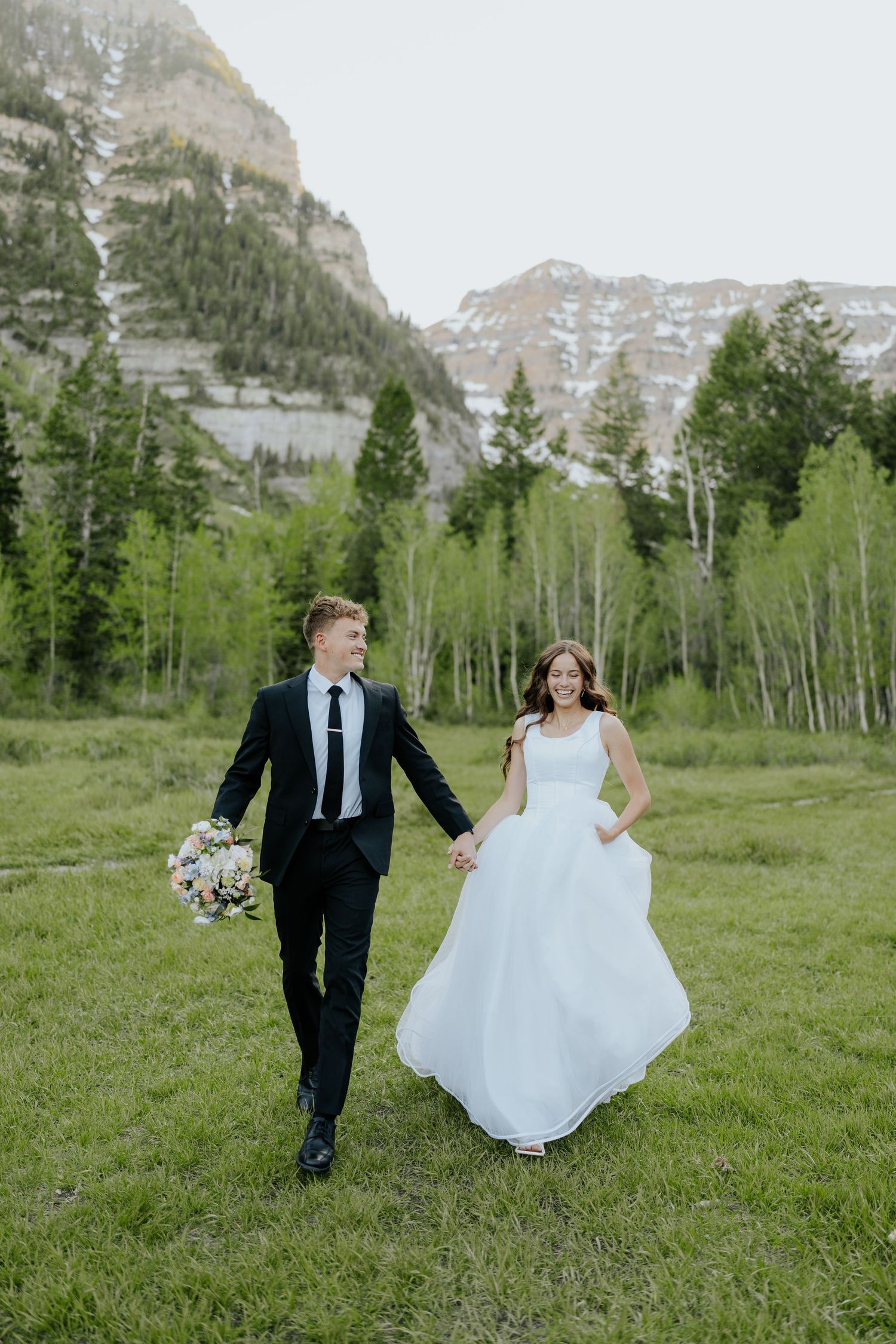 A smiling bride and groom walk hand-in-hand through a green mountain meadow, surrounded by aspen trees and rocky peaks.