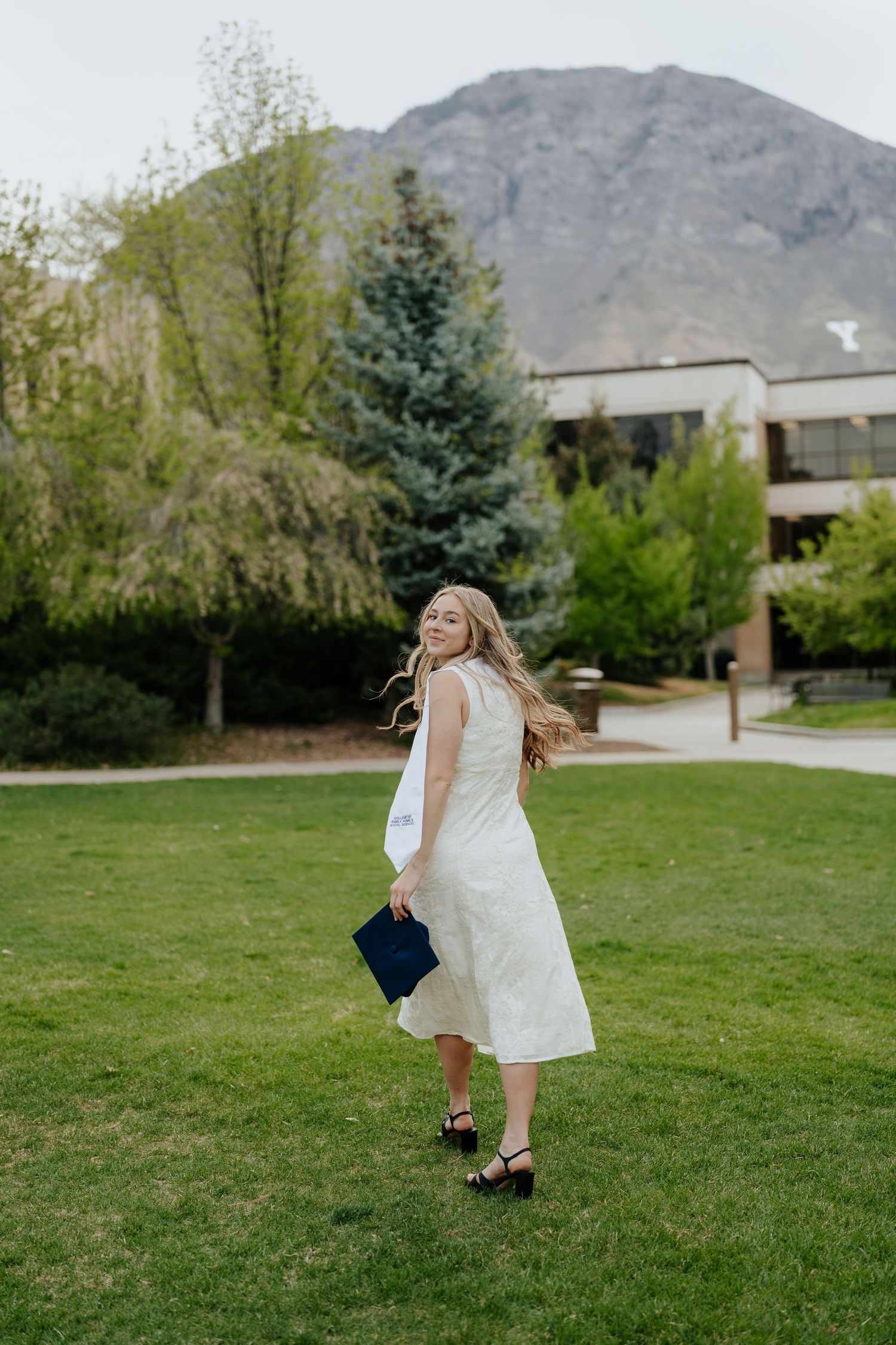 BYU Graduate in white midi dress twirling on green lawn with mountain scenery behind her.