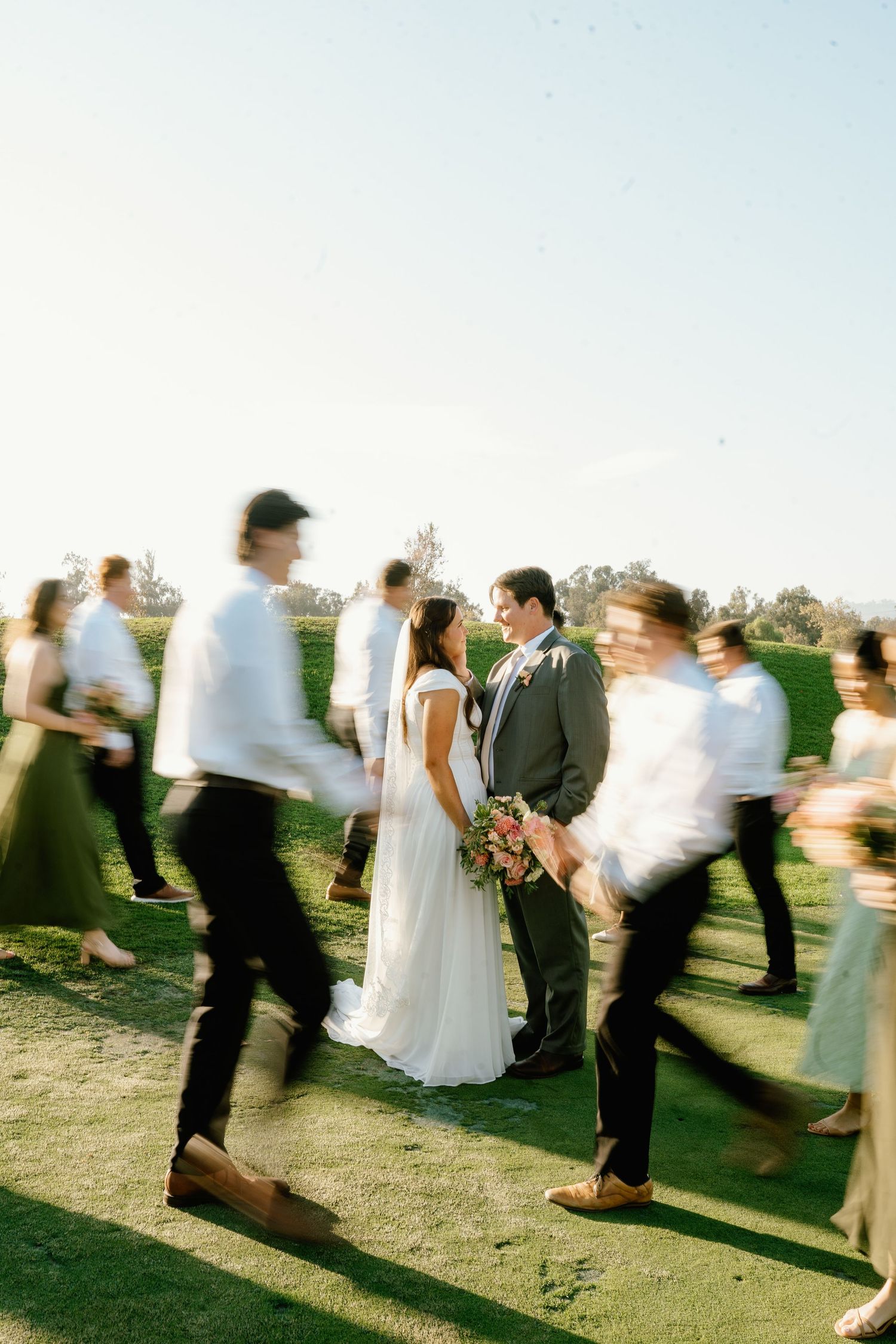 Bride and groom stand still as wedding guests blur around them on a sunny green lawn.