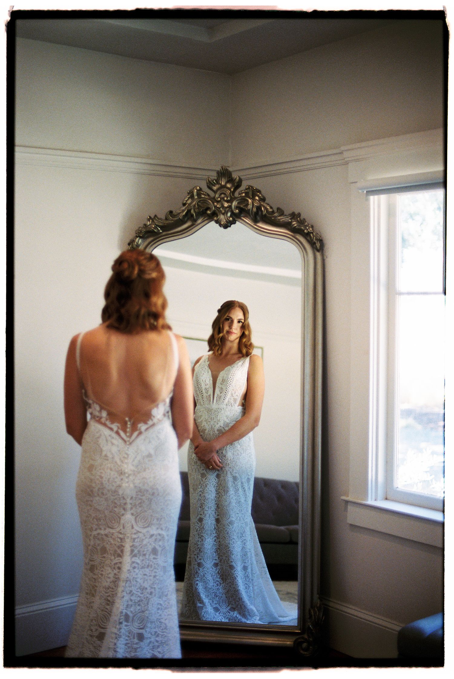 Bride in lace backless wedding gown admiring her reflection in an ornate gold floor mirror in a bright white room.
