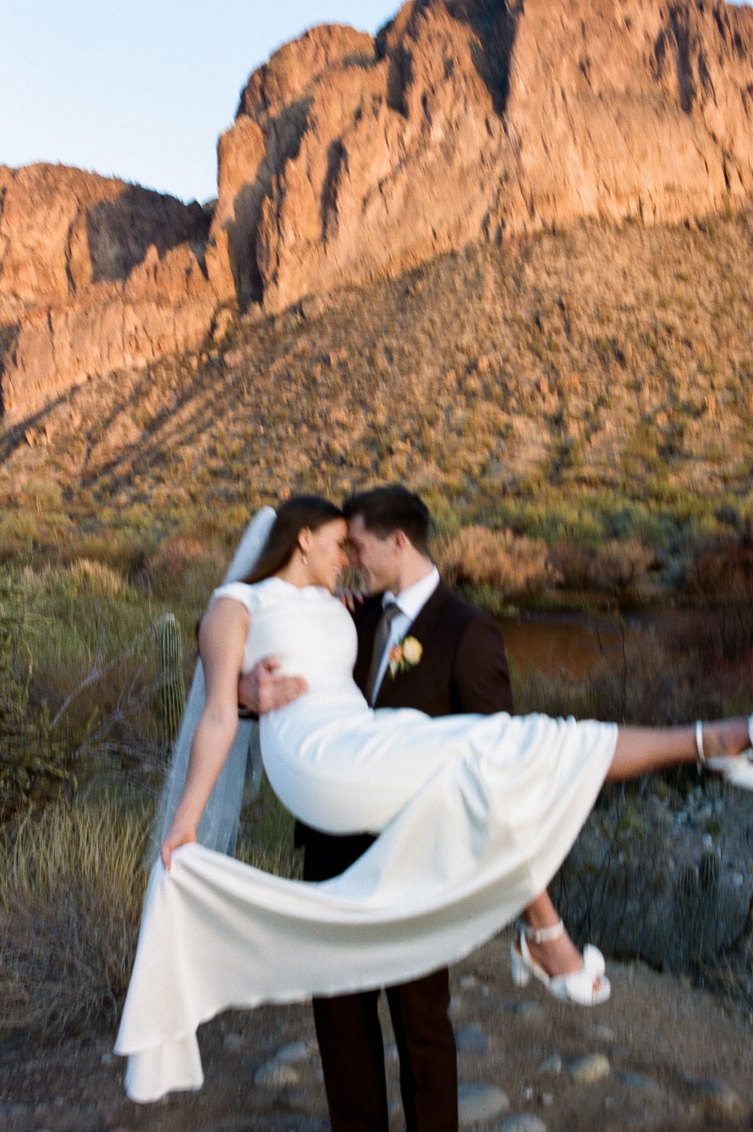 Groom lifts bride in wedding dress against dramatic red rock mountain landscape at golden hour.