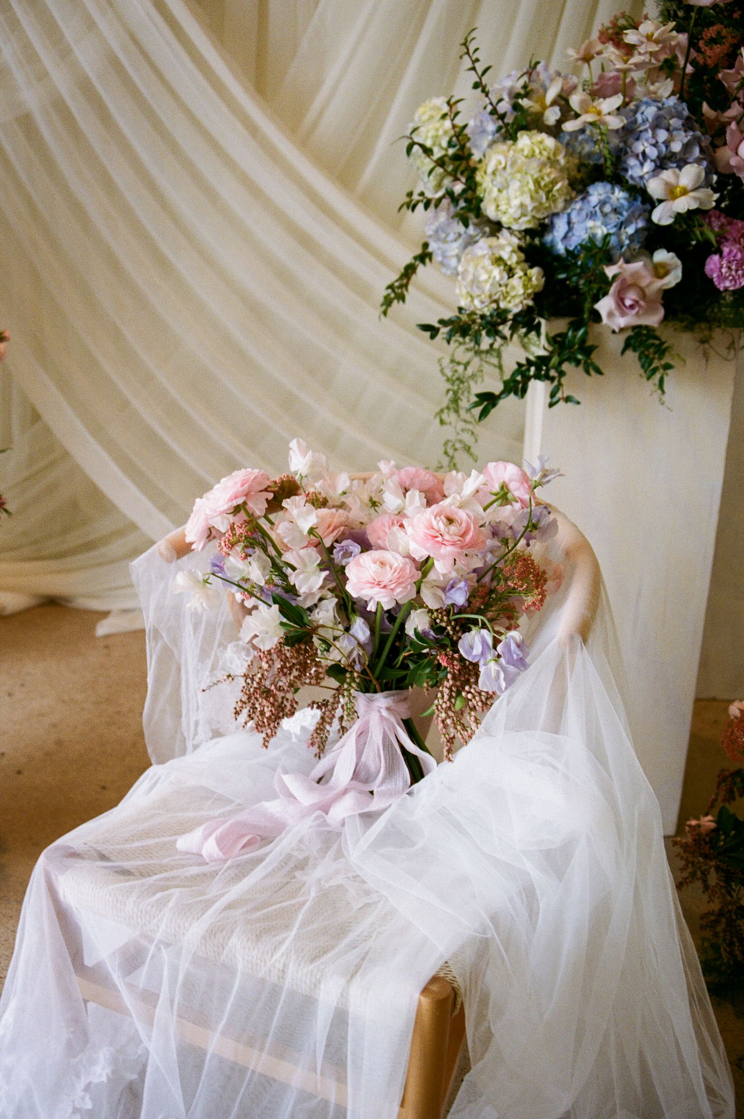 Romantic pink and white wedding bouquet draped in tulle rests on a chair with floral backdrop decorations.