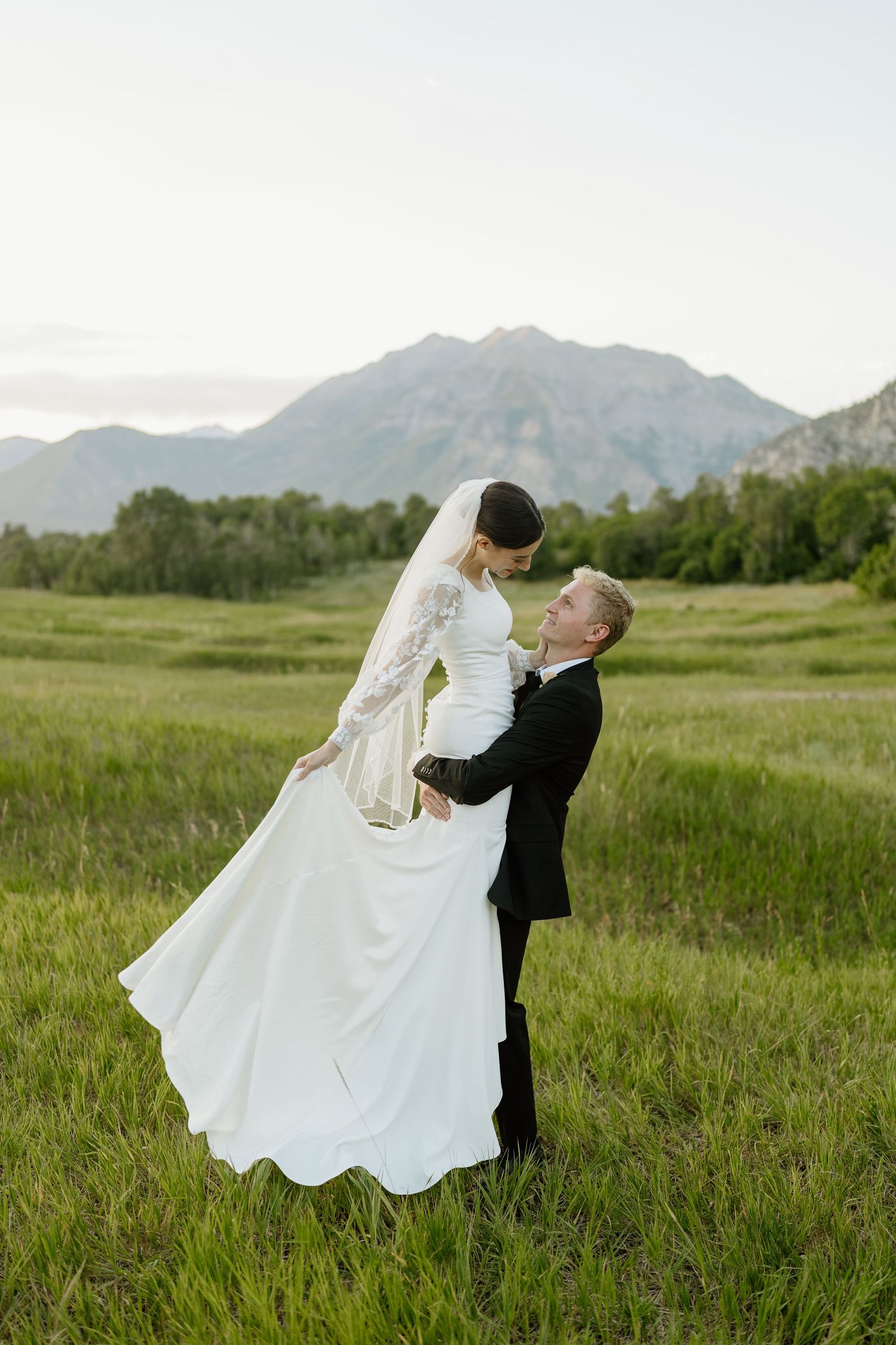 Groom lifting bride in white gown and veil in lush green meadow with mountain backdrop.