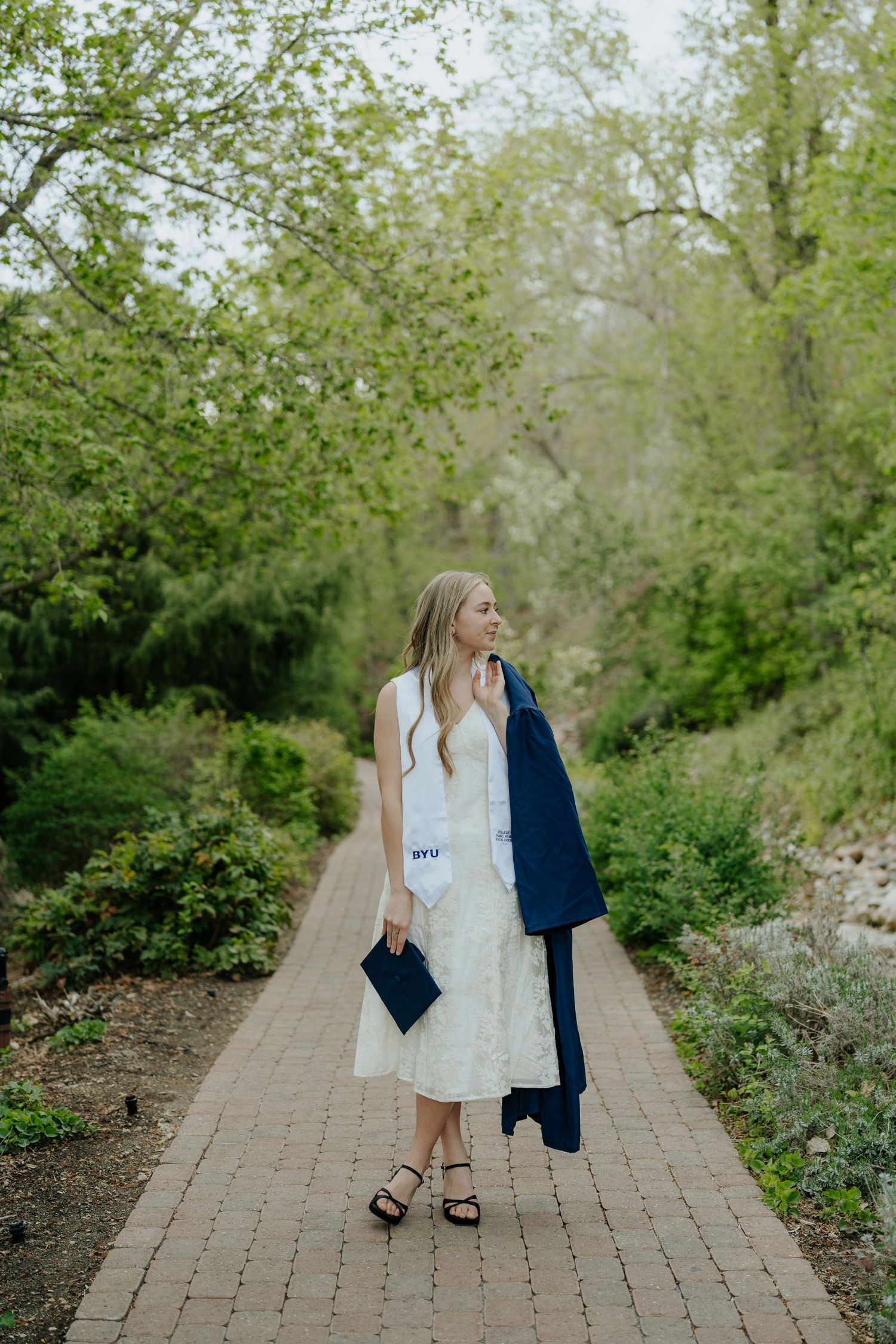 Blonde BYU graduate in white lace dress carrying navy gown on brick garden pathway.