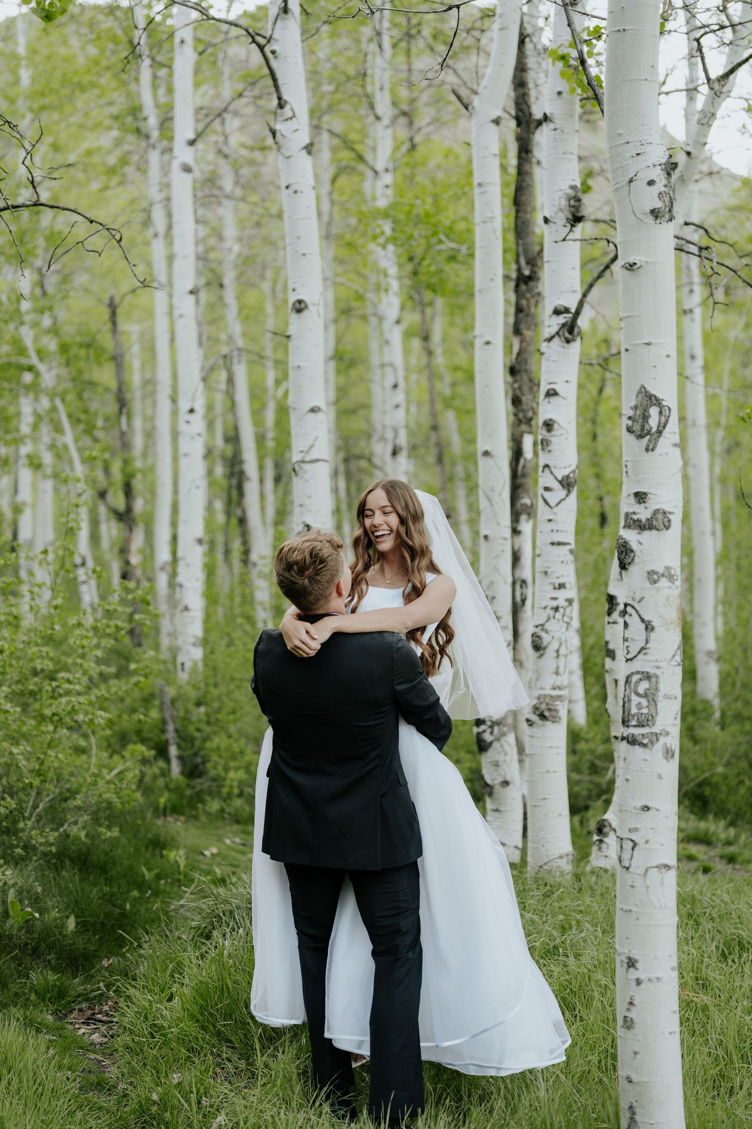Groom lifting bride in white gown among aspen trees in a lush green forest wedding photo.