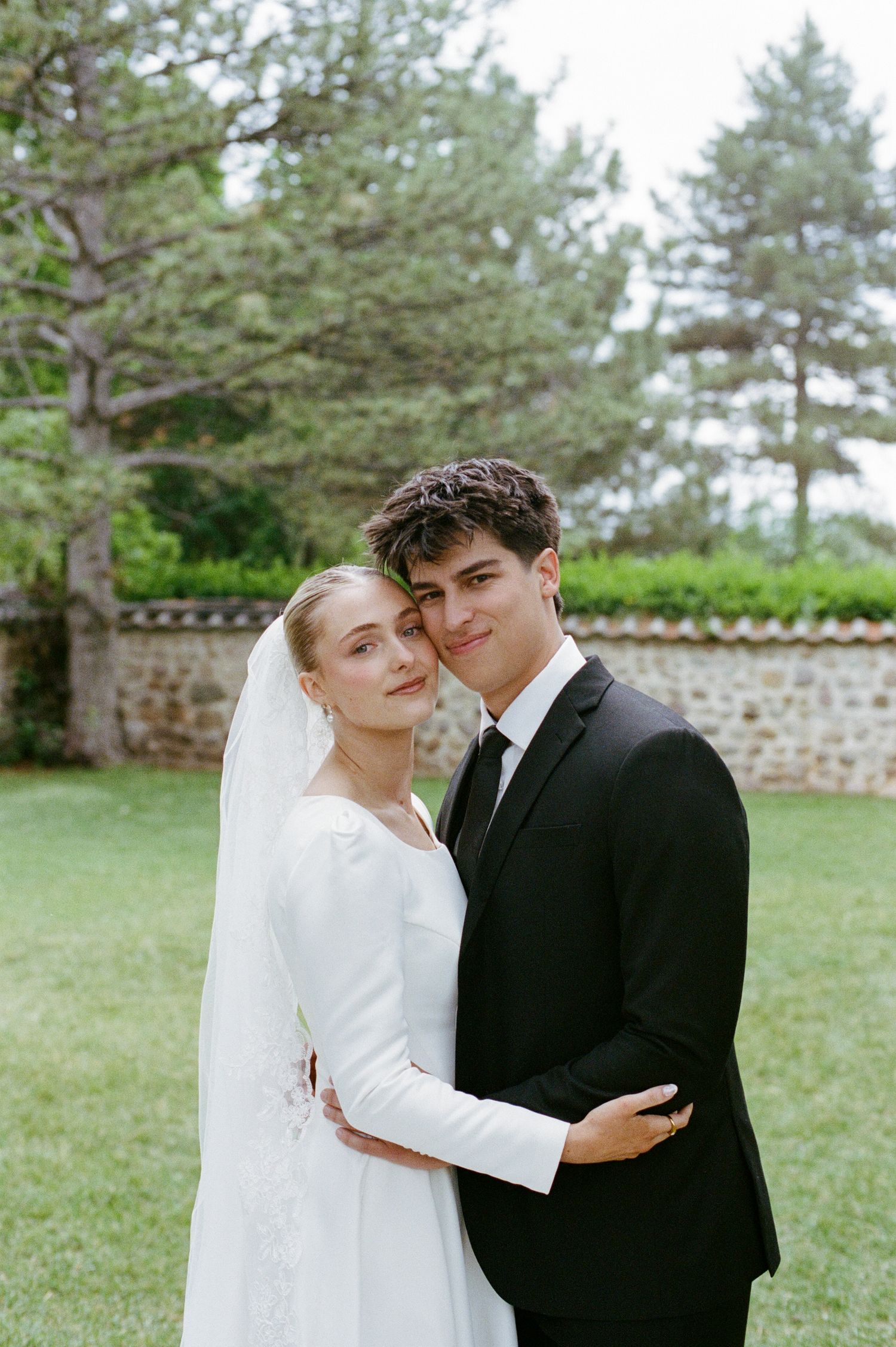 Bride in elegant white gown with veil embraces groom in black suit outdoors on lush green lawn.