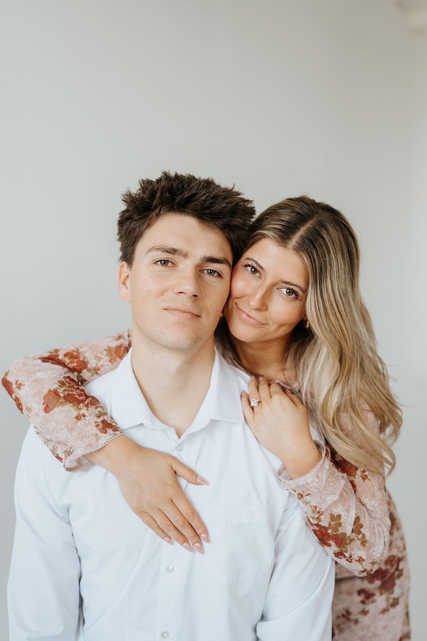 Couple posing together, woman in floral dress hugging man in white shirt against white background at Studio Elevn in Salt Lake City, Utah.