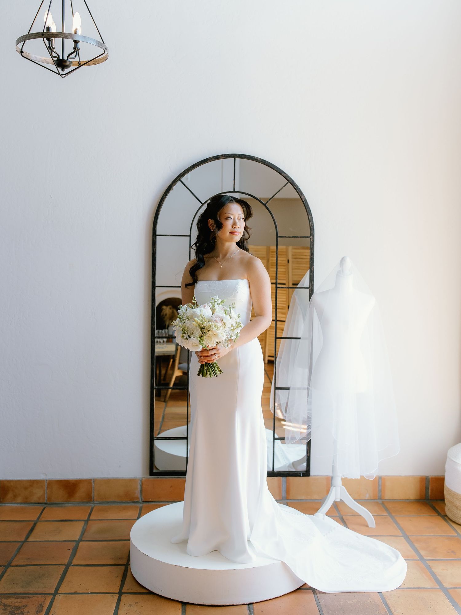 Bride in strapless white gown holds bouquet before arched mirror, with dress form visible in elegant bridal suite.