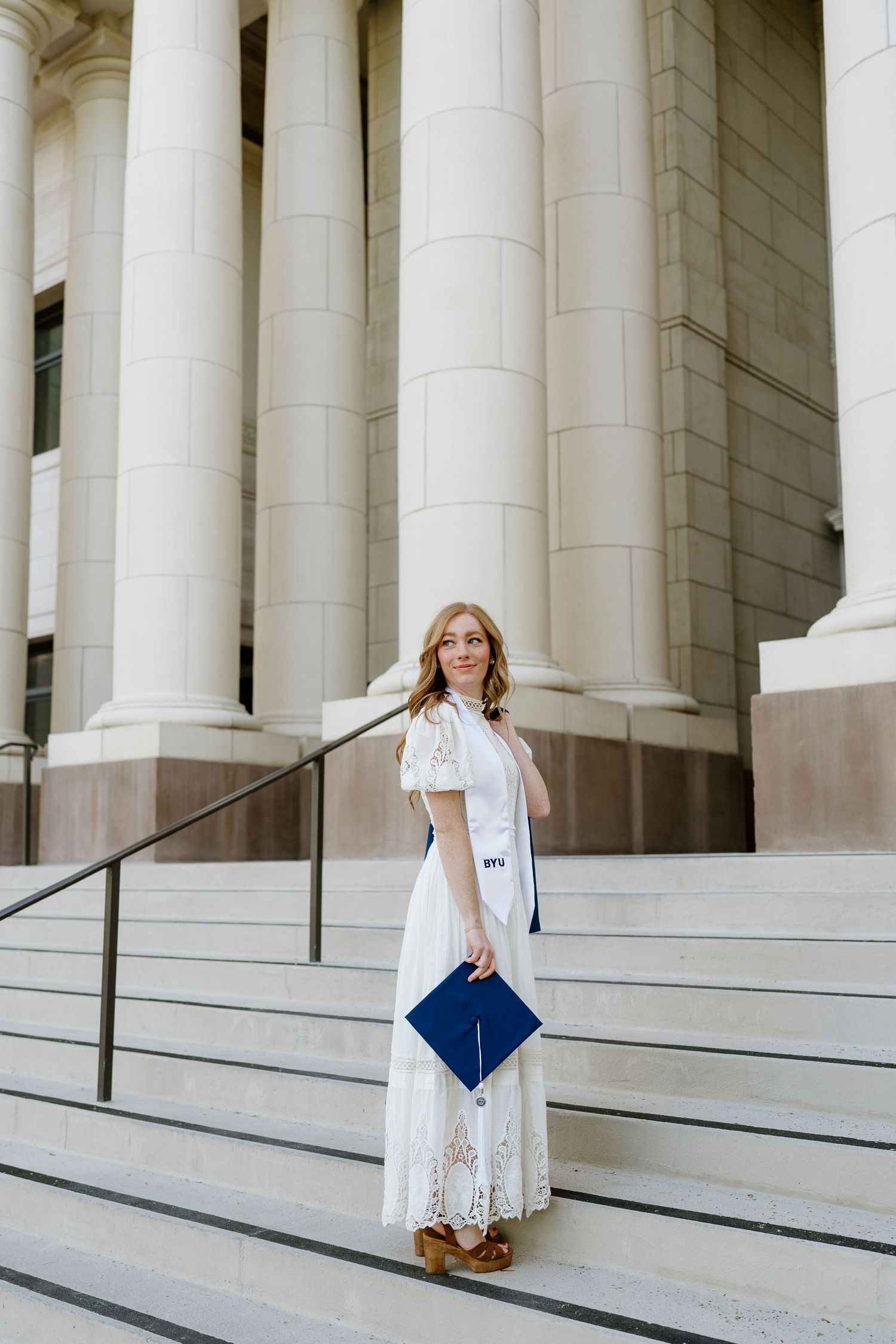 Blonde woman in white lace dress holding blue BYU graduation cap on stone steps.